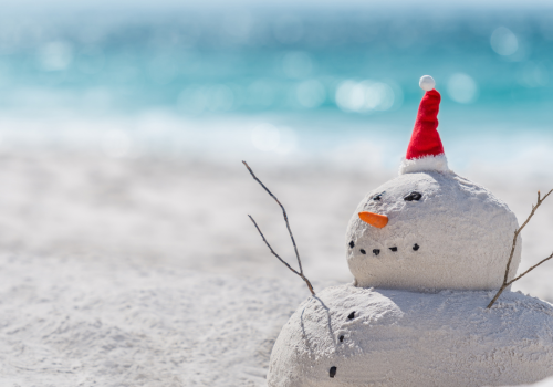 A sand snowman with a red Santa hat, sticks for arms, rocks for eyes, and a carrot nose is on a beach with the ocean in the background.