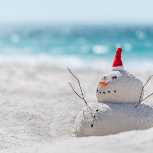 A sand snowman with a red Santa hat, sticks for arms, rocks for eyes, and a carrot nose is on a beach with the ocean in the background.