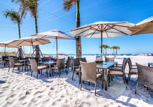 A beachfront restaurant with tables and chairs under umbrellas, set on white sand, with palm trees and a clear blue sky.