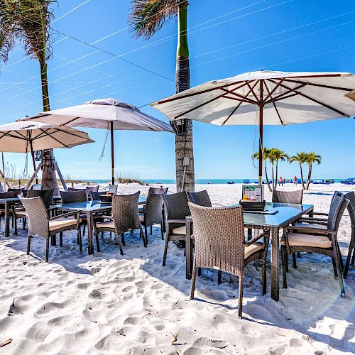 A beachfront restaurant with tables and chairs under umbrellas, set on white sand, with palm trees and a clear blue sky.