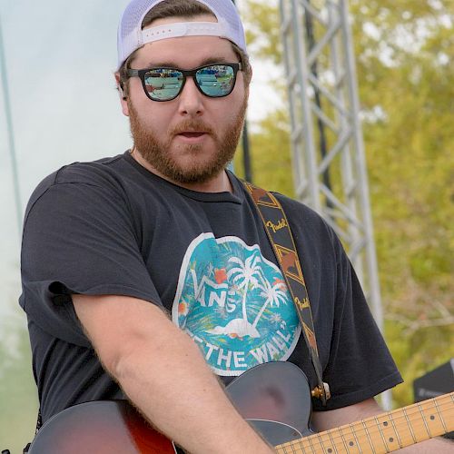 A person with sunglasses and a cap plays an electric guitar on stage at a Reggae Rise Up event, as indicated by the logo in the bottom corner.