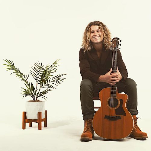 A person with long hair sits on a stool holding an acoustic guitar, with a potted plant next to them, against a plain background.