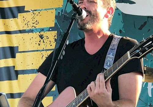 A man with a beard is singing and playing the guitar on an outdoor stage with a microphone stand in front of a colorful backdrop.