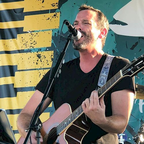 A man with a beard is singing and playing the guitar on an outdoor stage with a microphone stand in front of a colorful backdrop.