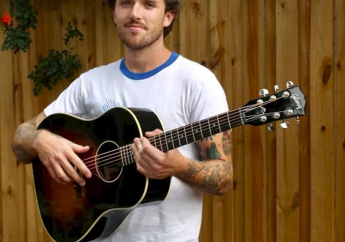 A man stands outdoors in front of a wooden fence, holding an acoustic guitar and wearing a white T-shirt and black pants.