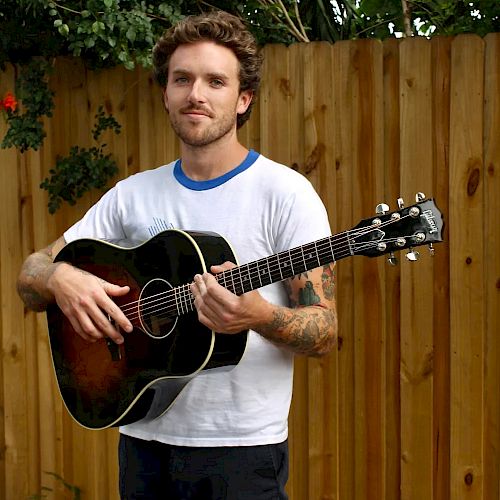 A man stands outdoors in front of a wooden fence, holding an acoustic guitar and wearing a white T-shirt and black pants.