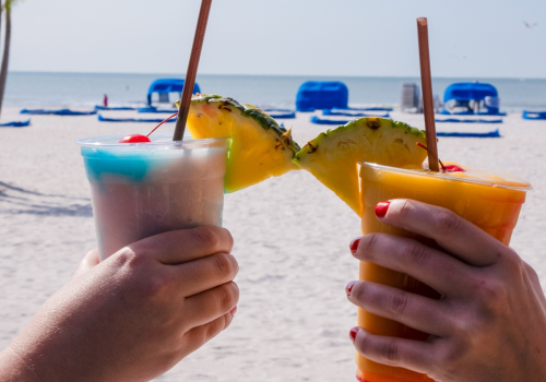 Two hands holding tropical drinks with pineapple garnishes near a beach with blue lounge chairs and the ocean in the background.