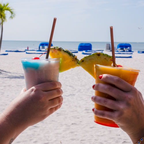 Two hands holding tropical drinks with pineapple garnishes near a beach with blue lounge chairs and the ocean in the background.