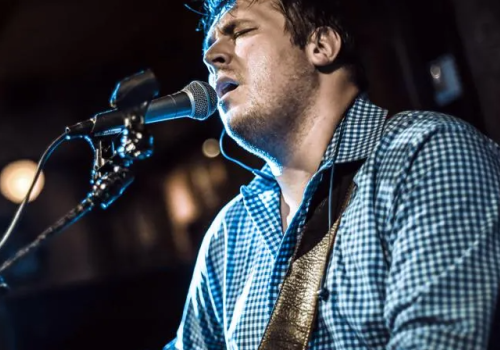 A man is passionately singing into a microphone while playing an acoustic guitar, dressed in a checkered shirt in a dimly lit setting.