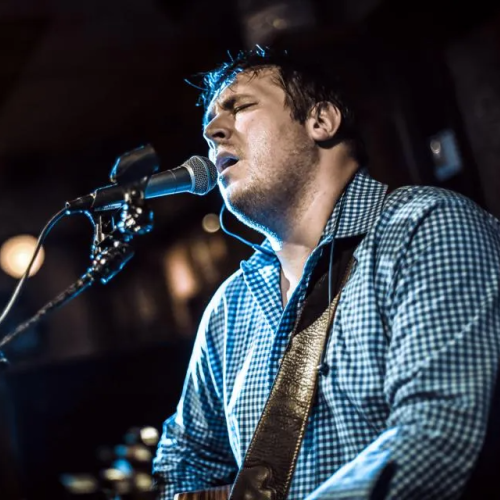 A man is passionately singing into a microphone while playing an acoustic guitar, dressed in a checkered shirt in a dimly lit setting.