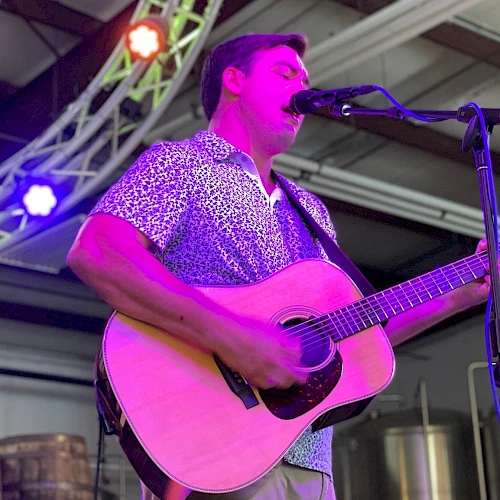 A musician plays an acoustic guitar and sings into a microphone on a stage with colorful lights overhead.