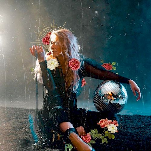 A person with long hair sits on the ground surrounded by flowers and a disco ball, with a mystical, ethereal vibe and a dark, misty background.