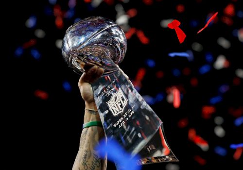 The image shows a person holding up the Vince Lombardi Trophy with confetti falling in the background, celebrating a Super Bowl victory.