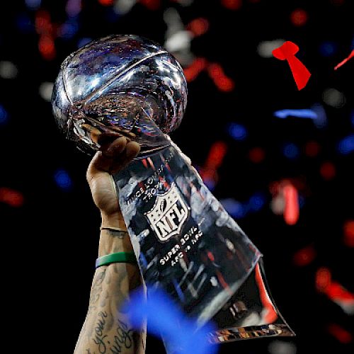 The image shows a person holding up the Vince Lombardi Trophy with confetti falling in the background, celebrating a Super Bowl victory.