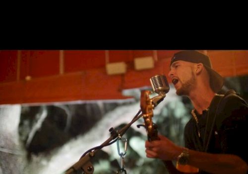 A person wearing a backward cap sings into a microphone while playing a guitar, possibly performing live.