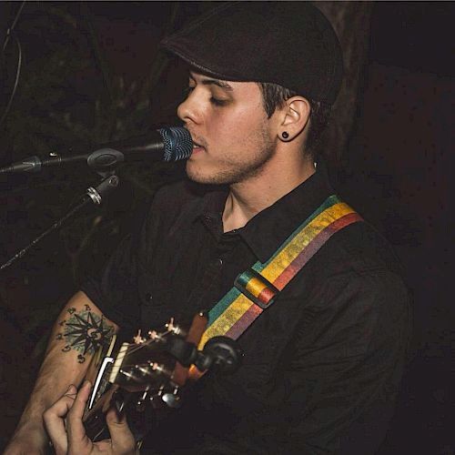 A person wearing a hat and black shirt is playing a guitar and singing into a microphone, with a colorful guitar strap around their shoulder.