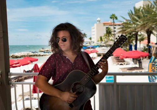 A person with long hair, wearing sunglasses and a red shirt, is playing a guitar on a beach with palm trees and red umbrellas in the background.