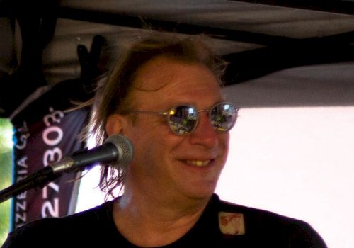 A person is on stage, playing guitar, wearing sunglasses, and a black t-shirt with a green peace symbol. The person is smiling.