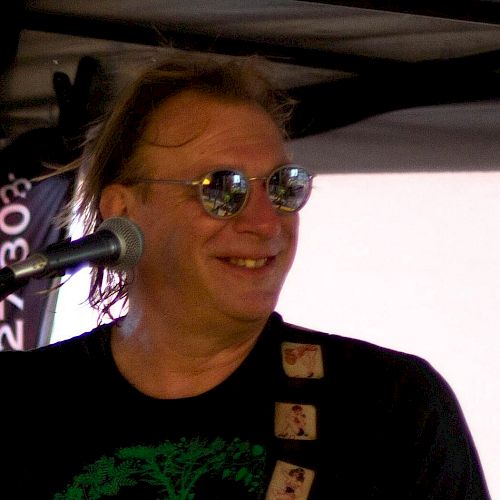 A person is on stage, playing guitar, wearing sunglasses, and a black t-shirt with a green peace symbol. The person is smiling.