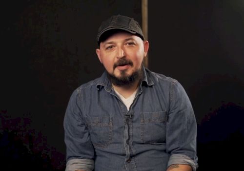 A person wearing a denim shirt and a cap is sitting against a dark background, looking towards the camera.