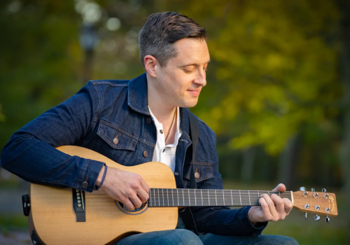 A person is sitting outside surrounded by greenery, playing an acoustic guitar while wearing a denim jacket and a white shirt, smiling slightly.