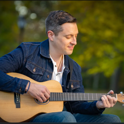 A person is sitting outside surrounded by greenery, playing an acoustic guitar while wearing a denim jacket and a white shirt, smiling slightly.