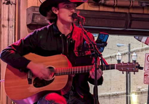 A musician in a cowboy hat performs with an acoustic guitar on a stool at a venue named 