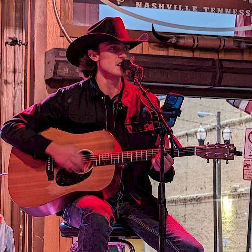 A musician in a cowboy hat performs with an acoustic guitar on a stool at a venue named 