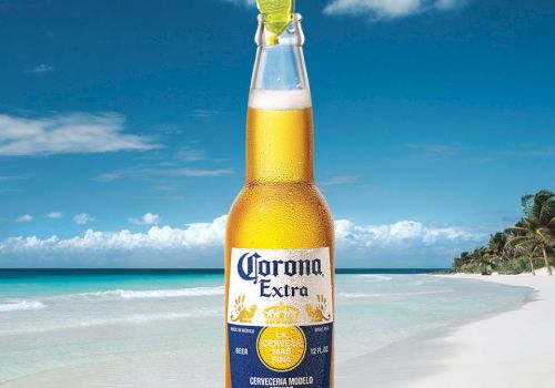 A bottle of Corona Extra beer with a lime wedge in the neck, set against a tropical beach background with clear blue skies and sandy shores.