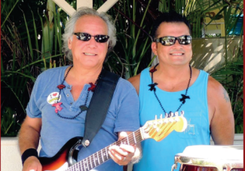 Two men are smiling, one holding an electric guitar while the other stands next to a conga drum. Both are wearing sunglasses, outdoors.