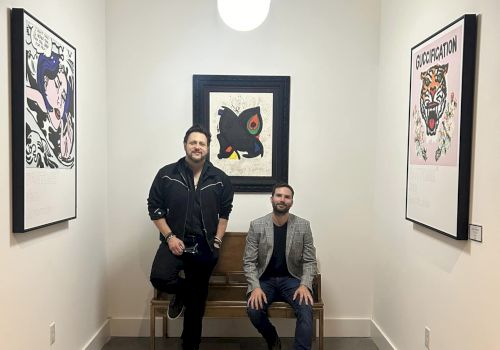 Two individuals in a small room with framed artwork on the walls; one standing, the other seated on a bench under a hanging light fixture.