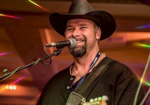 A person is singing and playing the guitar on stage, wearing a black hat and shirt. The background features colorful lights.