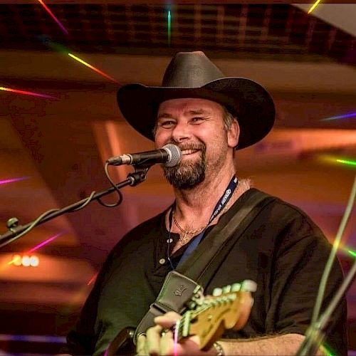 A person is singing and playing the guitar on stage, wearing a black hat and shirt. The background features colorful lights.