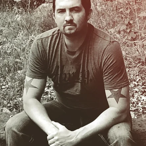 A man with star tattoos on his arms sits outdoors on a grassy and leafy ground, wearing a t-shirt and jeans, facing the camera with a serious expression.