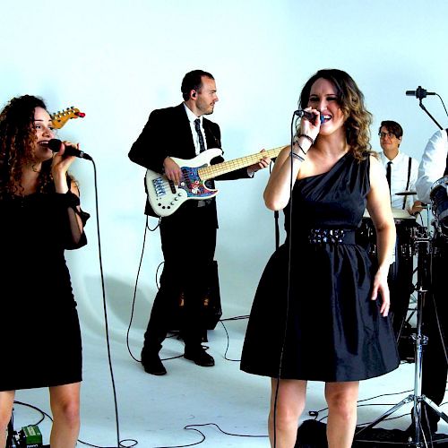 The image shows a band performing with two women singing, a man playing bass, and another man playing drums. They are in a bright studio setting.