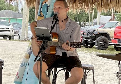 A person is playing an acoustic guitar and singing into a microphone under a thatched-roof structure, with vehicles and trees in the background.