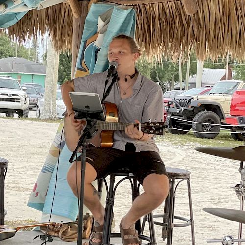 A person is playing an acoustic guitar and singing into a microphone under a thatched-roof structure, with vehicles and trees in the background.