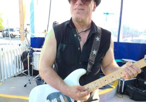 A man wearing a sleeveless shirt, hat, and sunglasses is playing an electric guitar on a stage. Drums are visible in the background.