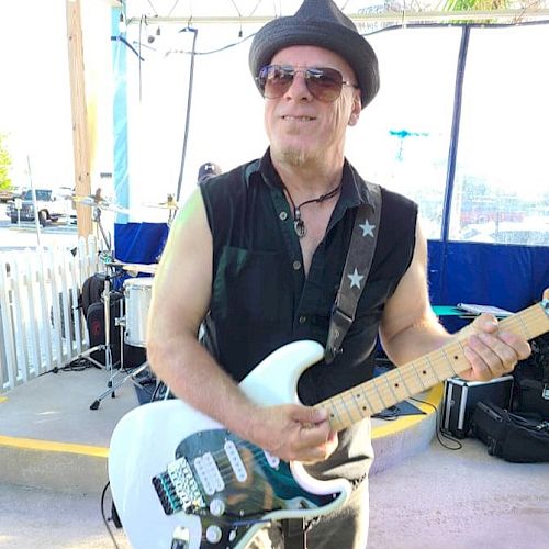 A man wearing a sleeveless shirt, hat, and sunglasses is playing an electric guitar on a stage. Drums are visible in the background.