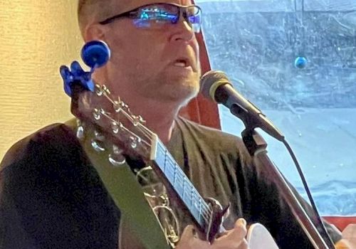 A person is singing into a microphone while playing an acoustic guitar. They are wearing glasses and appear to be performing indoors.