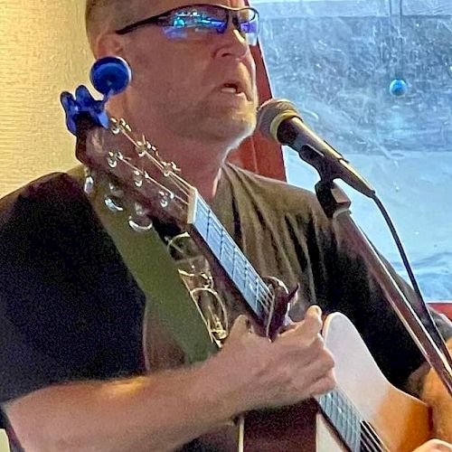 A person is singing into a microphone while playing an acoustic guitar. They are wearing glasses and appear to be performing indoors.