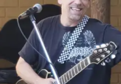 A man is smiling, holding an acoustic guitar, standing near a microphone. He is wearing a checkered hat and a black T-shirt with designs.