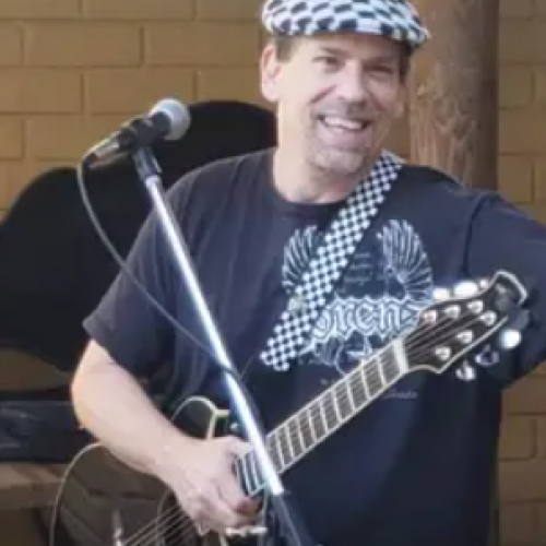 A man is smiling, holding an acoustic guitar, standing near a microphone. He is wearing a checkered hat and a black T-shirt with designs.