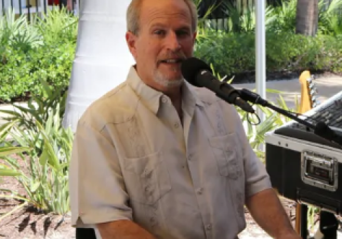 A man is playing a keyboard and singing into a microphone in an outdoor setting with greenery in the background.