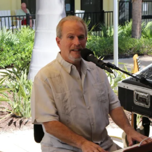 A man is playing a keyboard and singing into a microphone in an outdoor setting with greenery in the background.