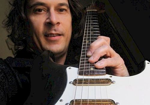 A person with long hair is holding a white electric guitar and smiling at the camera.