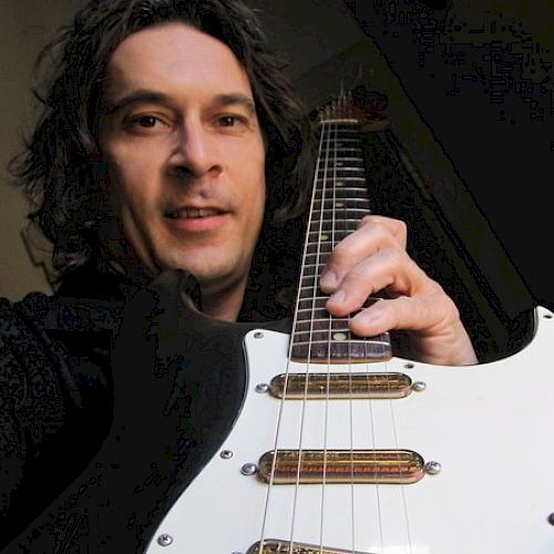 A person with long hair is holding a white electric guitar and smiling at the camera.