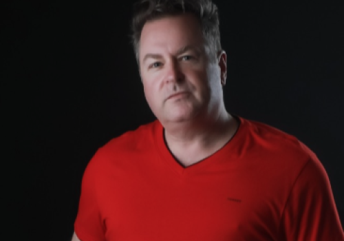 A person wearing a red shirt is posing in front of a dark background.