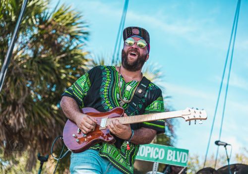 A person wearing a green-patterned shirt and cap is playing an electric guitar outdoors, next to a Topico Blvd sign, with palm trees in the background.