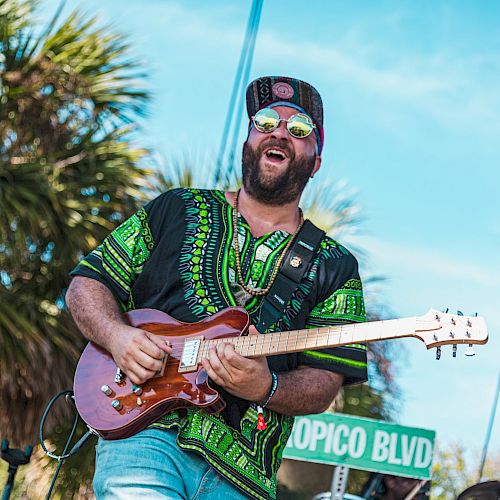 A person wearing a green-patterned shirt and cap is playing an electric guitar outdoors, next to a Topico Blvd sign, with palm trees in the background.